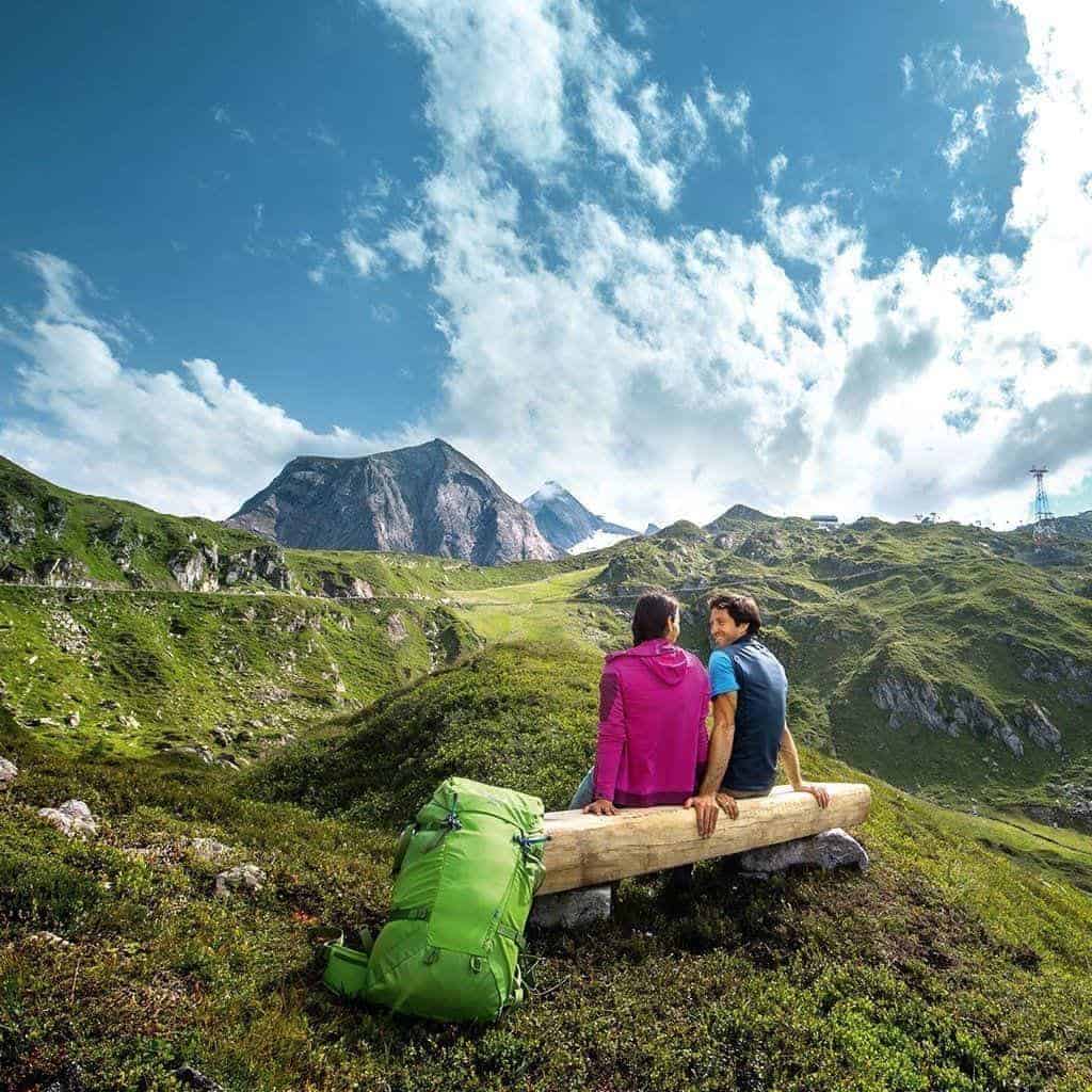 Paar sitzt auf einer Bank in den Bergen mit Blick auf die Alpenlandschaft und eine Seilbahn.